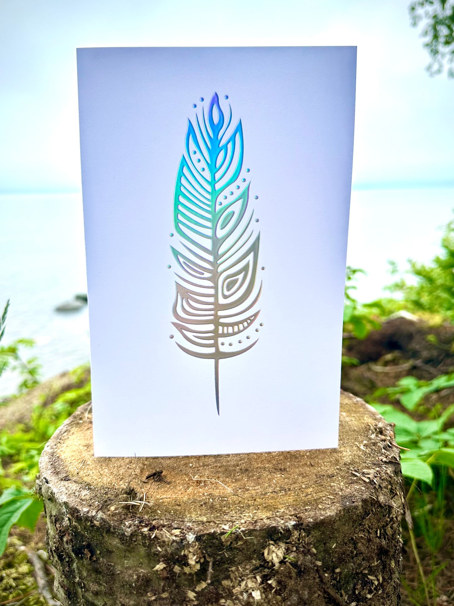 A greeting card with a woodland art style illustrated eagle feather. The feather has been treated with a holographic foil and reflects the light.